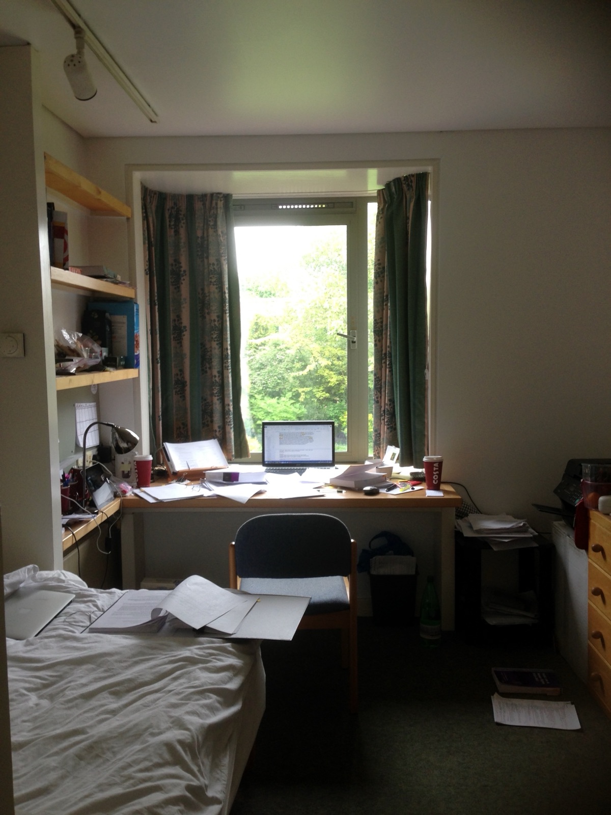 A desk under a window — a laptop mid-draft, stacked papers, two coffee cups, trees outside.